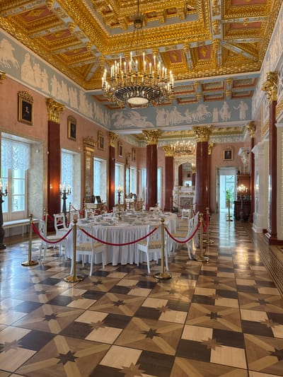 The image depicts a richly decorated interior, likely a banquet hall or grand room in a palace or historical building. The location is unknown. The room features a high, ornate ceiling with gilded details and painted frescoes. Large chandeliers hang from the ceiling. The walls are decorated with classical-style artwork and framed portraits. Red pillars and mirrored window placement line the wall on the right. A long table is set for a formal event, with white linens and chairs. Red rope barriers are placed around the table. The floor is covered in a parquet pattern. In the background, an open doorway leads to another room with plants.
