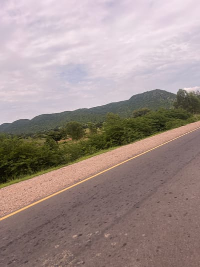 This media file shows a scenic view of a road running alongside a lush green mountainside in Minda, Nigeria. The road, paved with asphalt, has a distinct yellow line marking its edge, separating it from a gravel shoulder. The mountains, covered in dense vegetation, dominate the left side of the image, with their slopes stretching into the distance. A band of green trees and bushes runs along the base of the mountains, bordering the road. The sky above is overcast with light, wispy clouds, suggesting a diffused, possibly humid, atmosphere typical of this region. The scene appears to be captured during daylight hours, though the specific time of day is difficult to ascertain due to the cloudy conditions. No people or specific objects are prominently featured; the focus is on the natural landscape and the man-made road. The overall impression is one of a tranquil, rural setting, possibly a view from a moving vehicle.