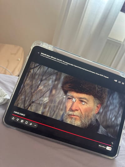 The image shows an iPad screen displaying a paused video of a movie titled "White Bim Black Ear" (translated), filmed in 1977 in the USSR. The video is from the channel "Old Soviet Films in HD Quality." The timecode shows 2:50:30 out of a total of 2:56:12. The iPad is encased in a clear plastic case and appears to be resting on a bed or similar soft surface. There is nothing in the image to suggest any location in the city of Gijón/Xixón, Spain.