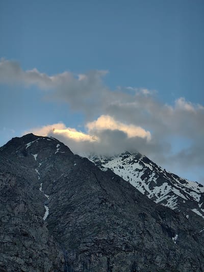 The image shows a majestic mountain range under a clear blue sky with scattered clouds. The mountains are rocky and rugged, with patches of snow near the peaks. The sunlight is hitting the top of one of the mountains, illuminating it with a warm glow and creating dramatic shadows on the slopes. Wispy clouds are catching the light, appearing golden and white against the blue sky. There are no people or objects visible in the image, and no text. The scene appears to be a natural landscape, likely captured during sunrise or sunset due to the lighting. The overall mood is serene and awe-inspiring, highlighting the grandeur of nature. The location is identified as Kead, India.