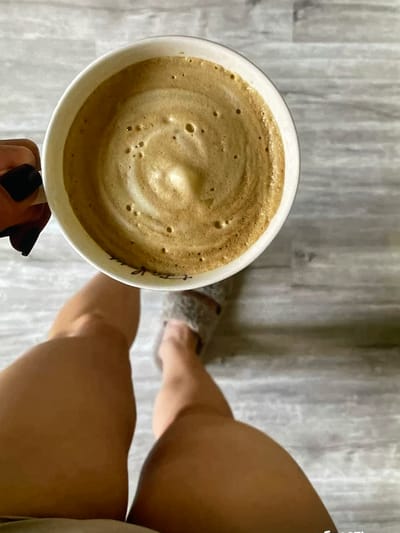 A top-down shot shows a white ceramic mug filled with a foamy coffee drink. The coffee has a swirl pattern on top. A hand with black nail polish is holding the mug. In the background, a person's legs are visible, wearing a fuzzy slipper on one foot. The scene appears to be set on a grey, wood-planked floor. There is no identifiable information or context to place this image in Neubrandenburg, Germany.