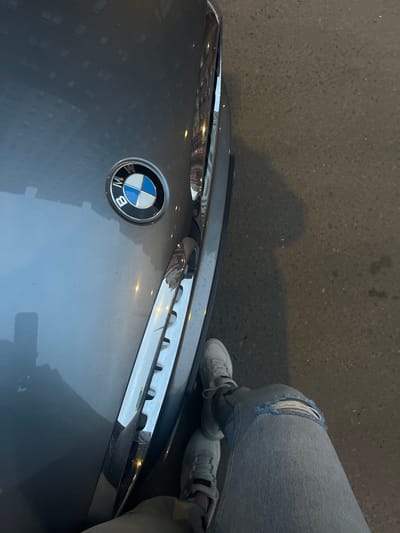 A BMW emblem is visible on the hood of a gray car. A person wearing ripped jeans and gray sneakers stands next to the car. The location appears to be an urban area, possibly in the city of Razvilka, Russia, although specific landmarks are not visible. The car's hood reflects the surrounding buildings.