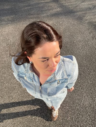 An overhead-view photograph depicts a solitary adult female standing on dark grey asphalt pavement in Montigny-le-Bretonneux, France. The subject has dark brown hair, parted on the side, with some strands appearing windblown. She is dressed in a light blue denim zippered dress or jumpsuit featuring a collar and chest pockets with metallic buttons. A silver-toned stud earring is visible on her right ear, and a silver-toned wristwatch is present on her left wrist. Her footwear consists of brown suede low-top sneakers with white stripes and white laces. Her head is angled downwards and to the right, with her gaze directed similarly. Direct sunlight illuminates the scene from the upper right, casting a distinct, elongated shadow of the subject to her left on the granular asphalt. An additional, elongated shadow from an off-frame object is visible on the asphalt in the upper left background.