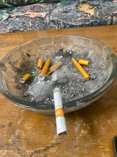 The image displays an indoor scene in Cimenyan, Indonesia, focusing on smoking paraphernalia. A clear, possibly glass, octagonal ashtray, approximately 20-25 cm in diameter, occupies the central mid-ground. The ashtray contains a dense accumulation of grey and black cigarette ash, alongside at least five discarded cigarette butts with distinctive yellow-orange filters. Remnants of a green-labeled material are also embedded within the ash. A faint plume of white smoke rises from the ash within the ashtray, suggesting recent activity.
In the immediate foreground, resting on a light-to-medium brown wooden table surface, is a single white cigarette. This cigarette features an orange and green band near its filter, with partially visible branding that appears to be "DIA MAN." The tip of this cigarette is burnt, and a small quantity of white ash has fallen onto the wooden surface directly beneath it. The wooden table exhibits a visible grain pattern and minor surface wear.
The background is blurred, revealing a piece of furniture, likely a sofa or armchair, upholstered with a multi-patterned fabric. The fabric displays a complex design incorporating shades of blue, green, red, and beige. A prominent worn area or tear is visible on the upholstery, exposing lighter material underneath. No individuals are present within the frame.