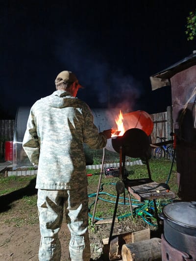 An outdoor nighttime scene in Kauchuk Stantsiya, Russia, features a person viewed from the back, wearing a full military-style digital camouflage uniform and a beige baseball cap. The individual is tending to a large, repurposed metal barrel serving as a barbecue grill or fire pit, which contains a bright, roaring fire, casting an orange glow.

The setting appears to be a rural backyard or similar outdoor area. To the left, a long, clear, semi-circular structure, resembling a greenhouse or storage tunnel, is visible, with a red barrel positioned behind it. A wooden fence runs across the mid-ground. On the right side of the image, part of a rustic wooden building or shed is seen, with a blue bicycle leaning against the fence.

The ground consists of a mix of dirt and patchy grass, and a green hose is snaked across it. Various grilling tools, including a spatula and tongs, are laid out on a small shelf attached to the grill stand. In the right foreground, a large dark pot with a lid sits on an outdoor stove or burner, with logs resting on the ground nearby.

The primary light source is the intense fire, which illuminates the immediate area and the person's front. Additionally, an artificial light source shines from behind the person, highlighting their back and the foreground, creating distinct shadows against the dark night sky. No discernible emotions are visible, and there is no visible text in the image. The activity suggests preparation for cooking or simply managing a campfire.