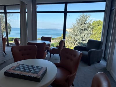 The image depicts a lounge area in what appears to be a hotel or resort, possibly in Bozhurets, Bulgaria, given the presence of the sea in the background and the general aesthetic. The foreground features several brown leather chairs arranged around white circular tables. On one of the tables is a decorative box with a chessboard design on top. Behind the seating area, large windows offer a panoramic view of the sea, with a distant coastline visible across the water. Trees and greenery are visible outside the window on the right side. A dark gray armchair is positioned near the window, facing away from the camera. White curtains hang on either side of the windows. The room is brightly lit with natural light. The flooring appears to be carpeted in a neutral gray tone. There are no people visible in the image.