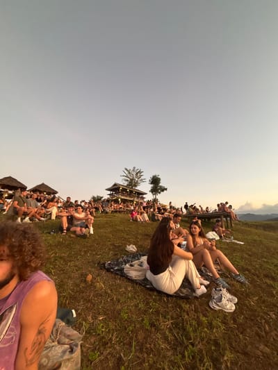The scene depicts a large outdoor social gathering on a grassy hillside in Ban Mae Ping, Thailand, at what appears to be late afternoon or sunset, indicated by the sky's hue transitioning from light blue to a warmer tone near the horizon. In the immediate foreground, to the left, is the partial profile of an individual with curly brown hair and a beard, wearing a purple tank top, seated on a patterned blanket. A tattoo is visible on their right upper arm. In the mid-ground, slightly to the right, two females are seated on a dark patterned blanket on the grass. The female on the left has long dark hair, is wearing a dark top and light-colored striped trousers, and is looking towards the second female. The second female, on the right, has brown hair, is wearing a white top and shorts, and is holding a small white object. A pair of white sneakers and a pair of blue and white sneakers are on the grass beside them.
Further up the hillside, a crowd estimated at several dozen individuals, primarily adults, are distributed across the slope. Many are seated directly on the grass or on blankets. To the left, several groups are gathered near thatched-roof structures. Centered towards the upper part of the hill is a prominent multi-story building, constructed in a traditional Thai architectural style with multiple balconies and a dark-tiled roof, surrounded by people socializing. Additional individuals are seated on elevated wooden platforms or decks to the right of the main building. Distant mountains or hills are visible on the horizon to the far right. The overall environment is open and illuminated by natural light, suggesting an informal event or gathering.