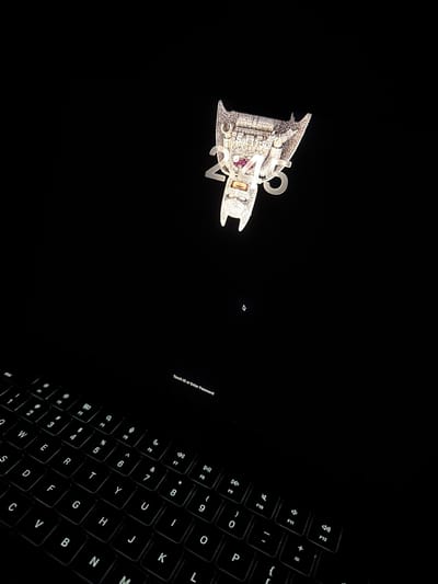 A close-up, high-angle shot captures a laptop screen and keyboard, likely in Vaughan, Canada. The screen displays a lock screen interface with a digital clock showing "2:45" above a prominent diamond-encrusted pendant resembling a winged creature, possibly a bat, with a red gem accent. The text "Touch ID or Enter Password" is visible in the lower right quadrant of the screen. Below the screen, the illuminated keyboard of the laptop is visible, with its keys showing a standard QWERTY layout. The background is entirely black, emphasizing the illuminated elements.