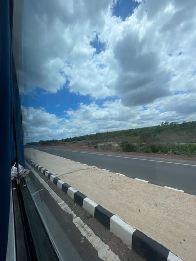 This image captures a view from inside a vehicle, looking out towards a highway in Sugali Tanda, India. The sky is filled with a mix of white and gray clouds against a bright blue backdrop. A paved road stretches into the distance, bordered by a rough shoulder and a concrete barrier with alternating black and white markings. Beyond the road, the landscape is covered in dense green vegetation and scattered rocks. The left side of the image shows the interior of the vehicle, including blue fabric upholstery and reflections on the window. There are no people visible in the scene.