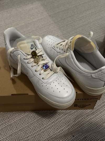 Two white low-top sneakers are displayed in an indoor, still-life setting, likely during the evening or night under artificial light, in Hainburg an der Donau, Austria. The shoes are placed on a brown cardboard box, partially visible.
The shoe in the foreground, angled towards the viewer, features two decorative rings adorning its off-white laces. One ring is rectangular with a prominent blue gemstone, while the other is an oval-shaped green/gold gemstone, positioned closer to the shoe's tongue. A "NIKE" logo is visible on the cream-colored tongue of this shoe. The shoe has small perforations on its toe box.
The second sneaker is positioned behind and slightly to the left of the first, with its sole facing the viewer. Both shoes appear to be made of white leather with light-colored soles and textured accents around the ankle collar.
The cardboard box beneath the shoes displays branding for "Dr. Martens," including "AIR WAIR" and "MARTENS.COM" text. The background consists of a light-colored, textured fabric, possibly a carpet or blanket, providing a soft contrast to the footwear. There are no people or discernible activities taking place beyond the arrangement of the shoes for display.
