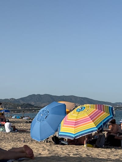 This is a photograph taken on the beach in Badalona, Spain, on a clear day. In the foreground, the sandy beach is populated with various beachgoers. Two umbrellas are prominently displayed: a solid blue umbrella to the left and a brightly colored striped umbrella to the right, featuring yellow, pink, orange, blue, and green stripes. Several people are visible in the background, some sitting on the sand and others under umbrellas. Further in the distance, a body of water, likely the Mediterranean Sea, is visible with a distant shoreline and hills under a pale blue sky. A bridge or pier structure can be seen spanning the water in the middle ground. The overall scene depicts a typical summer day at the beach.
