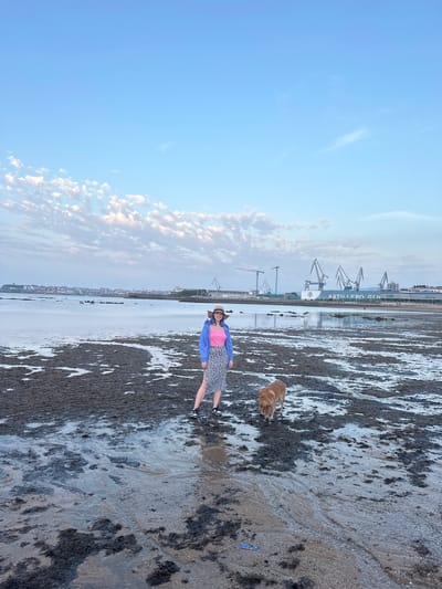 The image depicts a scene on a beach, likely at low tide, in Gijón/Xixón, Spain. A woman wearing a straw hat, blue jacket, pink top, and a patterned skirt is standing on the beach. A dog, likely a golden retriever, is nearby. The beach is covered in a mixture of sand and dark mud or algae. In the background, there is a line of industrial cranes and buildings, and the words "ASTILLERO GI" are visible. The sky is blue with some scattered clouds. The composition is vertical, with the beach stretching into the distance.
