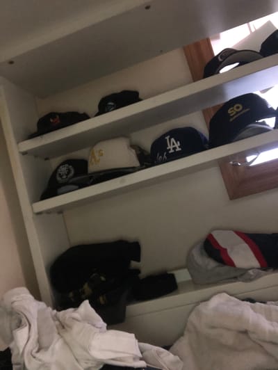 The image shows a shelf filled with baseball caps. The shelves are made of white material and appear to be part of a closet or storage unit. On the top shelf, there are two black caps, one with a red and yellow logo and another with a blue and white logo. The middle shelf has three caps: a black cap with a white logo, a white cap with a yellow logo, and a dark blue cap with a white "LA" logo. The bottom shelf has a black object that appears to be a backpack or large bag, and on the shelf above that, there's a gray and white striped item that looks like a folded blanket or pillow. The scene is indoors and dimly lit. The location is L'Hospitalet de Llobregat, Spain.