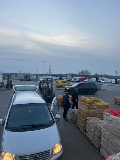 The image portrays an outdoor commercial loading area or market situated in Plodovitovo, Bulgaria, under an overcast sky consistent with either early morning or late afternoon. In the foreground, a silver Volkswagen Sharan-type minivan is parked, displaying illuminated headlights, an open rear hatch, and an open front passenger door. Adjacent to the minivan, to the right, two adult males are positioned on a paved surface. They stand near multiple palletized stacks of produce, primarily consisting of yellow mesh bags filled with potatoes, alongside additional stacks of grey and red mesh bags. One male wears a black hooded jacket and track pants with white side stripes. The second male, wearing a black jacket and a dark beanie, has orange gloves on both hands and appears to be interacting with the first male.
In the midground, a black Mercedes-Benz SUV is observed in motion, passing the individuals and produce stacks. Further back, a yellow Mercedes-Benz Sprinter van is visible. The surrounding area contains several other parked vehicles, including various vans and cars. Multiple industrial forklifts are strategically placed, suggesting ongoing or recent cargo handling operations. In the background, buildings resembling commercial structures or a gas station are discernible, along with numerous streetlights, power lines, utility poles, and sparse, bare trees. The ground throughout the visible area is composed of grey interlocking pavers.