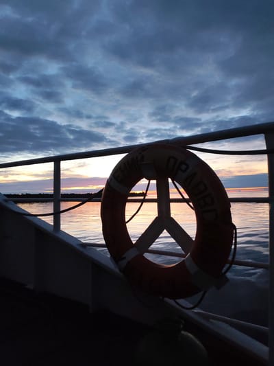 The image shows a lifebuoy attached to a railing on a boat. The boat is on a body of water, likely a river or lake, reflecting the colors of a sunset. The sky is filled with dramatic clouds. The lifebuoy is orange with white stripes and has Cyrillic lettering on it. The scene is set at dusk, with the low light casting long shadows. The location is identified as Shcheleiki, Russia.