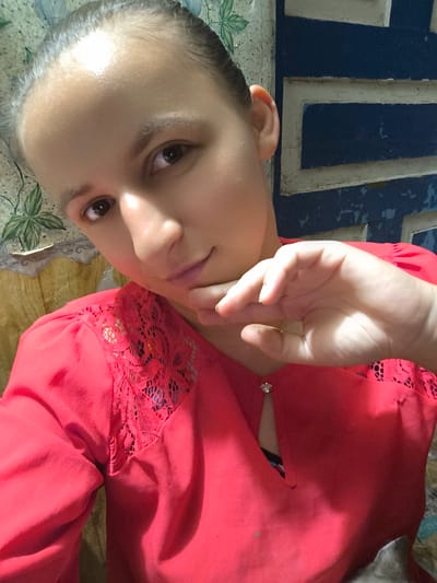 This is a close-up, selfie-style photograph of a young woman. She is the central subject, positioned in the foreground and filling most of the frame. The woman has her hair pulled back and is looking towards the camera. Her right hand is resting on her chin, a gesture that suggests thoughtfulness or contemplation. She is wearing a vibrant red top with lace detailing on the shoulders and a small, sparkly embellishment at the neckline.
The background is somewhat out of focus but reveals hints of the setting. To the left, there appears to be a floral-patterned curtain or wallpaper. To the right, a dark blue surface with lighter blue and white rectangular details is visible, possibly a painted wall or piece of furniture. The lighting suggests an indoor setting, likely during the day, with bright light reflecting off the woman's forehead.
There are no discernible objects other than the woman's clothing and hand. The activity is passive, with the woman posing for the photograph. No text is visible in the image. The overall mood is casual and personal, typical of a selfie. The image does not provide explicit clues about the time of day or weather conditions beyond the indoor setting. The woman's expression is neutral to slightly pleasant. The location context provided is Anastasivka, Ukraine.