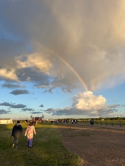 A wide-angle, eye-level shot captures a festival atmosphere in the city of Bolgar, Russia, under a dramatic sky. The late afternoon or early evening sun illuminates towering, fluffy clouds, casting a warm glow. A vibrant rainbow arcs across the sky, bridging the gap between the clouds and the horizon.

In the foreground, two individuals, likely a child and an adult, walk away from the camera across a grassy field. The adult, on the left, wears a dark jacket and jeans. The child, on the right, is dressed in a colorful floral jacket and purple pants. They are heading towards a distant stage setup with large screens and speakers, where a crowd of people is gathered. Tents, possibly vendor booths or event structures, are visible on the left side of the frame.

A black metal fence lines the right side of a gravel pathway, separating the festival grounds from a treeline in the distance. Several vehicles, including vans and possibly buses, are parked near the stage. The overall scene depicts a lively outdoor event with a natural spectacle overhead.