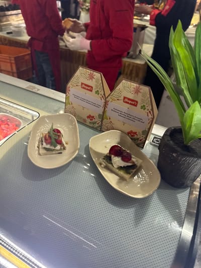 Two appetizer plates sit on a surface, each containing a small square of food topped with white cream and red toppings. Beside them are two Ganpati's boxes, one describing the contents as "Fettuennta, Strawberry Avocado, Hazelnut, Ricotta, Basil, Olive Oil and balsamic reduction", the other with an unreadable description. A plant in a dark gray pot sits behind the boxes. A person in red with pink gloves is visible in the background, holding a piece of bread or pastry.