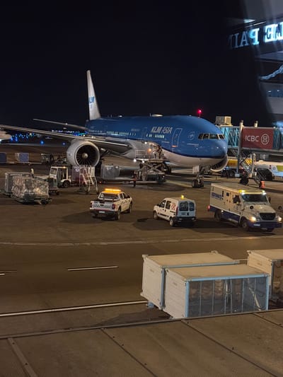 At night, at Aeropuerto Internacional Ezeiza in Argentina, a KLM Asia airplane is parked at the gate. The airplane is blue and white with "KLM ASIA" written on its side. A jet bridge connects the airplane to a terminal building, which has illuminated signs. In front of the airplane, there are various ground support vehicles, including baggage carts, a tow tractor, and a white van with orange flashing lights. A Brinks armored truck is also present. Several people are visible on the tarmac, attending to the aircraft and its cargo. The scene is illuminated by artificial lights from the terminal and vehicles, creating a well-lit tarmac.