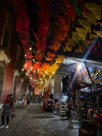 The image shows a nighttime street market in Oaxaca City, Mexico. A narrow alleyway is crowded with vendors selling colorful textiles, handicrafts, and souvenirs. Strings of lights and papel picado (perforated paper banners) illuminate the scene. People are walking through the market. Buildings line the sides of the street.