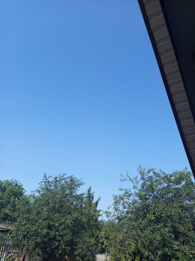 The image depicts a clear blue sky above a dense canopy of green trees. A portion of a building's soffit is visible in the upper right corner, angled downwards and to the left. The trees appear to be located in an outdoor, potentially rural or suburban, environment, consistent with a location like Хальчанскі сельскі Савет, Belarus. The scene is tranquil, with no people or specific activities readily apparent. The lighting suggests it is daytime, with the sun likely high in the sky.