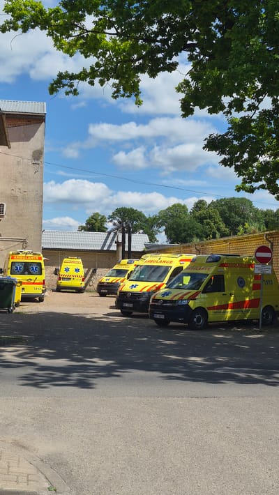 In Jēkabpils, Latvia, a row of five yellow ambulances are parked on asphalt outside a brick building. The ambulances are marked with "AMBULANCE" in red letters, along with a blue star emblem and the number "113". The ambulance closest to the right has a red and white circular sign with a horizontal red bar, indicating "No Entry", and the number "113" below it. The scene is illuminated by daylight, casting shadows from the trees onto the asphalt. The sky is blue with scattered white clouds.