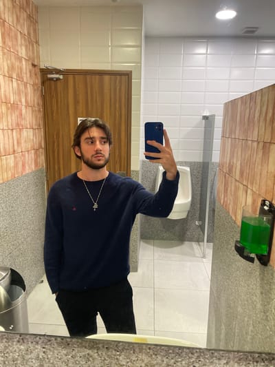 A young adult male is taking a mirror selfie in a public restroom. He is wearing a navy blue sweater and black pants, with a silver necklace featuring a cross pendant. The restroom has tiled walls, a wooden door, a urinal, a trash can, and a soap dispenser with green liquid. The image was taken in Cuautitlán Izcalli, Mexico.