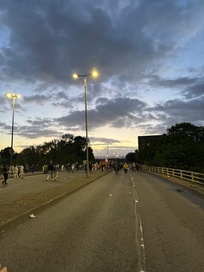 The image depicts a bridge over a road in Glasgow, United Kingdom, at dusk. The sky is filled with dramatic, dark clouds interspersed with patches of lighter blue and the warm glow of the setting sun near the horizon. Two tall lamp posts with multiple lights are visible, illuminating the scene. A procession of people is walking along the road, appearing to be moving from left to right in the frame. The road is asphalt, with white painted lines marking lanes. A wooden railing with yellow traffic cones lines the right side of the bridge. Trees and a modern building with illuminated windows are visible in the background on the right, while more trees and distant city lights can be seen on the left. The overall atmosphere suggests a public gathering or event taking place as evening approaches.