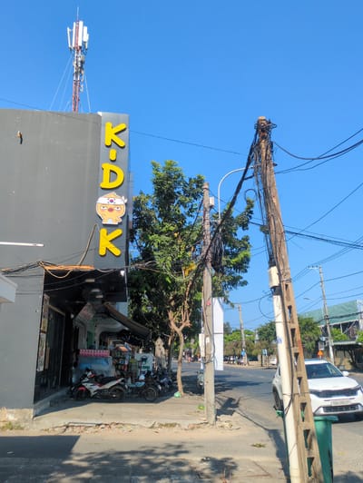 A street scene in Tuy Hòa, Vietnam features a gray building with a yellow "K-A-OK" sign and a character on its roof. A cell tower extends above the building. Power lines and utility poles are heavily bundled with wires. Several parked motorbikes are on the sidewalk in front of a commercial establishment. A white KIA car is parked on the street further down the road. The sky is clear and blue.