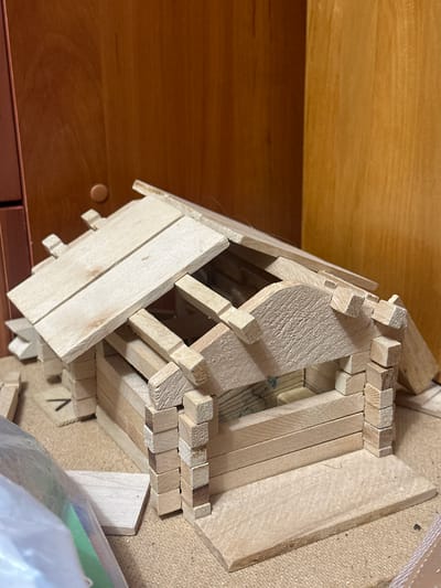 The image shows a partially constructed wooden model house. The house is made of small, unfinished wooden pieces of various shapes and sizes, including planks and blocks. The walls are constructed with stacked horizontal pieces and vertical corner supports. The roof is also made of wooden planks, some overlapping and angled. The model is resting on a textured, light brown surface, possibly burlap or a similar material. In the background, a wooden cabinet or wall with a circular knob is visible. There are also some loose wooden pieces and a clear plastic bag with colorful contents on the left side of the frame. The lighting suggests an indoor environment. The image does not contain any elements that indicate a specific location in Chornomors'ke, Ukraine.