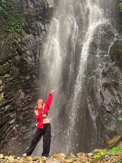 A full-body outdoor shot captures a young Caucasian woman standing on rocks at the base of a tall waterfall in Tu Le, Vietnam. The woman, with long blonde hair, is positioned slightly to the left of the frame's center, facing the camera with a smiling expression and her mouth partially open, with her left hand brought towards her mouth in a gesture, and her right arm raised with an open hand. She wears a dark olive-green crop top, a bright red long-sleeved jacket worn open and loosely over her shoulders, and baggy black cargo pants. On her feet are light-colored open-toed sandals. Her posture is dynamic, with her left leg extended forward and her right leg bent at the knee, giving the impression of movement or an active pose.
Behind her, the impressive waterfall dominates the scene, cascading down a rugged, dark grey rock face that is partially covered in lush green moss and small plants, particularly on the left side of the frame. The water flow appears strong and wide, creating a misty atmosphere. The rocks beneath the woman are irregular, angular, and vary in size and color, ranging from dark greys to lighter browns and oranges, suggesting a natural, untamed environment. Scattered green foliage is visible among the rocks in the lower right foreground. The lighting suggests an overcast or shaded condition, highlighting the water's movement and the textural details of the rocks.
