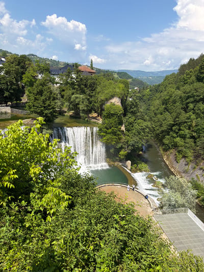 This is a vertical, high-angle shot of the Pliva Waterfall in Jajce, Bosnia and Herzegovina. The waterfall is the prominent subject, cascading down into a pool of green water. The scene is lush with dense green foliage and trees surrounding the waterfall and the river.
In the foreground, a thick cluster of vibrant green leaves dominates the right side of the frame. Behind the waterfall, a circular, stone-lined pool is visible, with a few people standing on the edge. Further upstream, a modern building with a translucent, diamond-patterned facade is situated next to the river. The river itself is seen flowing, with some white water indicating rapids.
Beyond the immediate waterfall area, the landscape opens up to reveal a town nestled on a hillside, with houses having tiled roofs. In the distance, rolling hills covered in trees recede into the horizon, under a bright blue sky with scattered white clouds.
The time of day appears to be daytime, likely late morning or early afternoon, given the bright sunlight and clear sky. The weather seems pleasant and sunny, ideal for outdoor viewing. The overall mood is serene and picturesque, highlighting the natural beauty of the location. There is no visible text in the image.