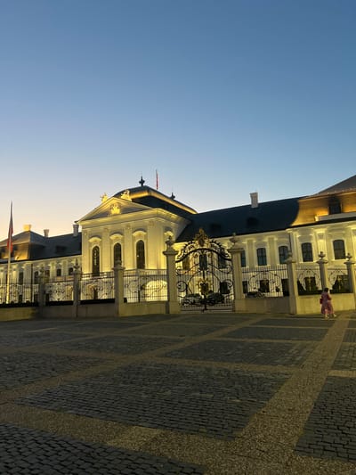 The image shows a building with a dark roof and a white facade, featuring arched windows and decorative elements. A wrought-iron gate with ornate scrollwork guards an entrance. The building is situated on a cobblestone plaza, with a pattern of lighter stone creating diagonal lines across the surface. The sky is clear and blue, transitioning to a lighter hue near the horizon, suggesting either dawn or dusk. A person wearing a pink garment is visible in the distance, walking away from the camera. Two flags are also present, one prominently displayed on a pole near the building's facade, and another smaller one near the bottom of the frame. The lighting indicates that the sun is low in the sky, casting long shadows and highlighting the architectural details of the building. The overall scene depicts a public space in front of a historical or government building, likely in Bratislava, Slovakia, given the location context. There is no visible text in the image.