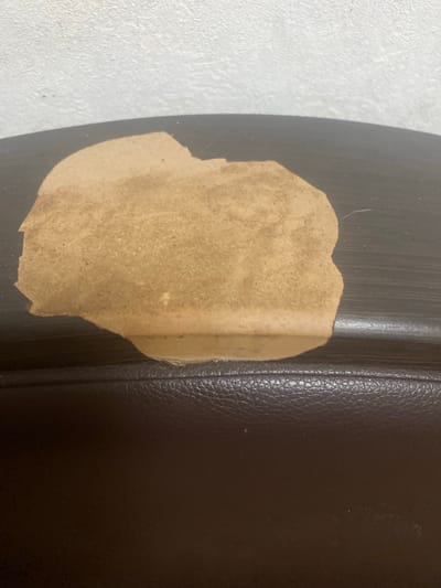 This image shows a close-up of a dark brown, textured surface that appears to be leather, possibly from furniture. A piece of light brown paper or sticker is affixed to the surface, obscuring a portion of it. The paper has an irregular shape with torn edges. The textured surface has stitching visible along the right side, indicating it might be a padded or upholstered item. The background to the left of the leather surface is a white, textured wall. The lighting suggests an indoor environment with a light source casting a shadow from the paper onto the leather. There are no people visible in the image, and no specific activity is taking place. The notable details are the contrast between the smooth, dark leather and the rough, light-colored paper, as well as the texture of both materials and the wall. No text is visible in the image.
