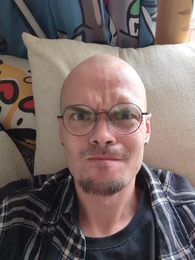 A close-up, rotated selfie depicts a bald Caucasian male wearing round-rimmed glasses and a dark blue and white plaid shirt. He has a goatee. He is lying down on a textured, beige pillow. Behind the pillow are colorful cushions, including one with a heart design and another with blue and green patterns. The location, Shunde District, China, cannot be verified based on the visual information alone.