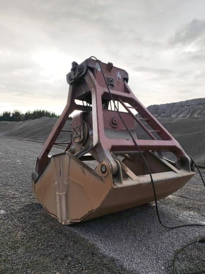 A rusty red industrial grabber bucket is positioned on a gravel surface. The grabber, a large piece of machinery, appears weathered and shows signs of use. Behind it, a vast pile of grey gravel extends into the distance. A line of trees can be seen on the horizon. The sky above is overcast with clouds. The scene suggests an outdoor industrial or quarrying environment. The image is likely taken in Shal'skii, Russia, a location known for its industrial activities related to construction materials.