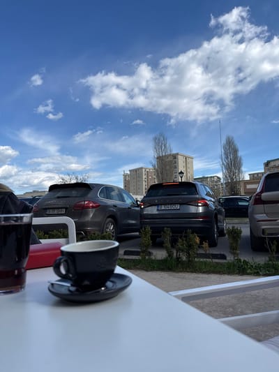 A low-angle, outdoor shot depicts a coffee cup and saucer sitting on a white table, alongside other indiscernible items. Several parked cars line the street, including a dark gray Skoda Kodiaq with license plate "B 104 GOU" and a gray Porsche Cayenne with license plate "B 111 SHH". High-rise buildings and trees dot the background under a cloudy blue sky. Based on the metadata, the location is Crețuleasca, Romania.