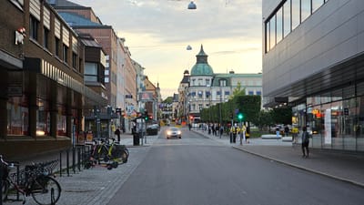 This media file captures a street scene in Linköping, Sweden, likely during the late afternoon or early evening as suggested by the soft, warm light and long shadows. The weather appears to be calm.
The scene depicts a moderately busy urban street lined with buildings on both sides. On the left, a brick building features storefronts with awnings and large windows, displaying illuminated advertisements. Text visible includes "pwc" on a sign and "ICA Supermarket" with "Öppet 7-22" (Open 7-22). There are bicycle racks with several parked bicycles and scooters in the foreground.
The right side of the street has a modern building with a reflective facade, obscuring the interior details. Further down the street, a more traditional European architectural style is evident, with a prominent building featuring a green dome. Trees line the sidewalk on this side, and people are walking and gathering. Traffic lights are visible, with a car driving down the street. Several individuals are walking on the sidewalks, some in groups. A few people are wearing yellow shirts.
The overall mood is tranquil and ordinary, portraying a typical street life in a Swedish city. The perspective is from the middle of the street, looking down its length towards the horizon where the sky is a soft blend of blue and warm hues, indicating dusk.