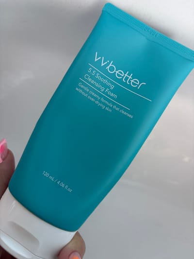 This is a close-up, angled shot of a turquoise tube of "vvbetter 5.5 Soothing Cleansing Foam" held by a person's hand, the lower part of which is visible on the right side of the frame. The tube is positioned diagonally from the top left to the bottom right of the image. White text on the tube indicates the brand name "vvbetter," "5.5 Soothing," "Cleansing Foam," "Gentle creamy formula that cleanses without over-drying skin," and the volume "120 mL / 4.06 fl.oz". There is also a small mark "DK004" near the top of the tube. The background is a plain, light-colored surface, likely a countertop or table, suggesting an indoor setting. The lighting is bright and even, indicating it could be daytime or well-lit indoor conditions. The focus is sharp on the product packaging and the hand holding it. There are no strong emotional cues, but the image appears to be a product showcase or personal photograph.