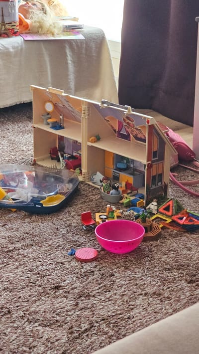 This media file captures a playful scene featuring a dollhouse and various toys scattered across a textured, brown carpet. The dollhouse, a prominent object, is detailed with multiple levels and rooms, suggesting it is a central element of the play. Inside and around the dollhouse, a collection of small figures and accessories are visible, including what appear to be people, animals, and furniture pieces.

Among the objects, a bright pink, bowl-shaped toy sits prominently, alongside other smaller toys like a red chair and a pink, textured disc. There's also a transparent plastic container filled with more toys. In the background, a glimpse of a doll with blonde hair is visible, adding to the sense of a child's play area. The overall impression is one of a casual, domestic setting, likely a child's playroom or living area, where imaginative play is actively taking place. The lighting suggests it could be daytime, as there is a natural light source illuminating the scene, possibly from a window. No specific weather conditions are discernible. No text is visible within the frame.
