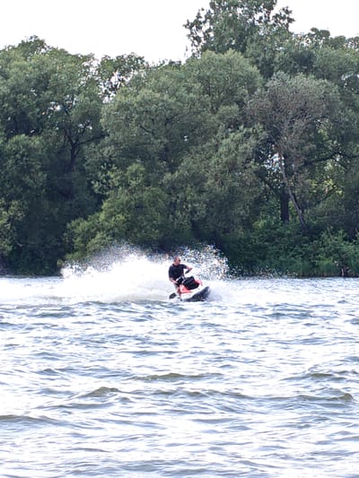 In the waters near Ждановіцкі сельскі Савет, Belarus, a person is riding a jet ski. The jet ski, which appears to be orange and black, is creating a large spray of water as it moves. The rider is wearing a black wetsuit and sunglasses. The surrounding environment is a body of water with choppy waves, and a dense line of green trees forms the background. The sky is overcast.