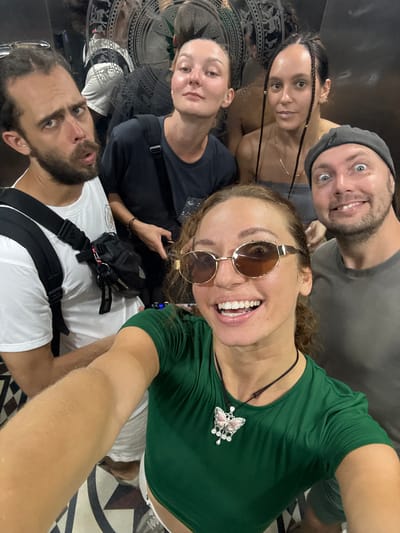 This is an upward-angled group selfie featuring five adults in a highly reflective indoor space, possibly an elevator or mirrored room.
In the foreground, a woman with curly brown hair, wearing round sunglasses and a deep green top, smiles widely, revealing her teeth. A prominent silver and pink butterfly necklace with clear stone accents adorns her chest. Her outstretched arm suggests she is holding the camera. Directly above her, a man with a short beard and a grey beanie smiles towards the lens. To the mid-left, a woman with braided dark hair looks at the camera with a calm expression.
In the bottom left of the frame, a bearded man wearing a white t-shirt and a black harness or backpack with an "adidas" logo visible on one strap looks upward with a slightly surprised or intense expression. Another woman, wearing a black t-shirt, is partially visible in the mid-left background, also looking towards the camera.
The reflective surfaces behind the subjects display distorted reflections of the individuals and a distinct circular pattern on what appears to be a floor or decorative panel. This ornate design features intricate geometric shapes and stylized animal figures, characteristic of traditional Southeast Asian art. The overall atmosphere is casual and cheerful, with most subjects smiling. The indoor lighting does not provide specific cues about the time of day or weather. The location context of Hà Giang 1 Ward, Vietnam, aligns with the potential cultural motifs seen in the background pattern.