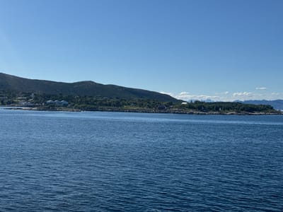 This is a scenic landscape photograph taken from the water, looking towards a coastal town in Bognes, Norway. The scene is dominated by a vast expanse of deep blue water in the foreground, with gentle ripples reflecting the sunlight. In the midground, a rocky coastline is dotted with buildings, including what appears to be a white church with a steeple, several houses, and larger industrial-looking structures. Lush green trees and vegetation cover the hills behind the town. The background features rolling, verdant hills that gradually recede into the distance, and further back, faint outlines of distant mountains can be seen under a clear, bright blue sky.
The time of day appears to be midday or early afternoon, given the bright sunlight and clear sky. The weather is fair and sunny, with no clouds visible over the water or the immediate coastline, though some wispy clouds are present near the distant mountains. The overall mood is peaceful and serene, showcasing the natural beauty of the Norwegian coast. There are no people or specific activities clearly depicted, the focus is on the tranquil landscape and the coastal settlement. No visible text is present in the image.