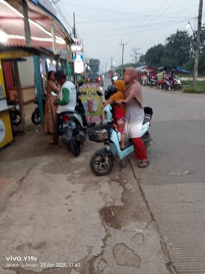 The image shows a street scene in Kecamatan Kroya, Indonesia, with two motorcycles parked at the side of the road. A woman in a brown headscarf and a red patterned dress is seated on a light blue electric motorcycle with a small child seated in front of her. The woman is wearing red sandals. Another woman is standing beside the motorcycle. A man in a green shirt is standing next to a black motorcycle. A woman in a long brown dress is standing in the background. The road surface is gray asphalt and the area is paved with concrete. There are power lines running overhead. The scene is partly obscured by a yellow building on the left of the frame with a sign in Indonesian language. A sign with the words "Teh Cing" is visible behind the motorcycles. The photo was taken on 23 April 2025 at 17:43. There is an empty space on the sidewalk in front of the motorcycles.