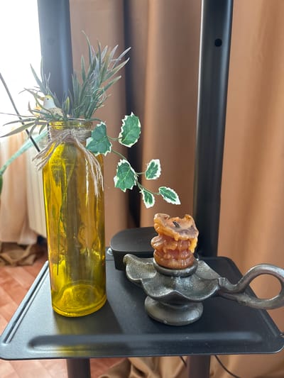 A yellow glass bottle with greenery and a small bird figurine is placed on a black shelf next to a burnt candle in a metal candlestick. The background consists of a brown curtain. The scene is indoors.