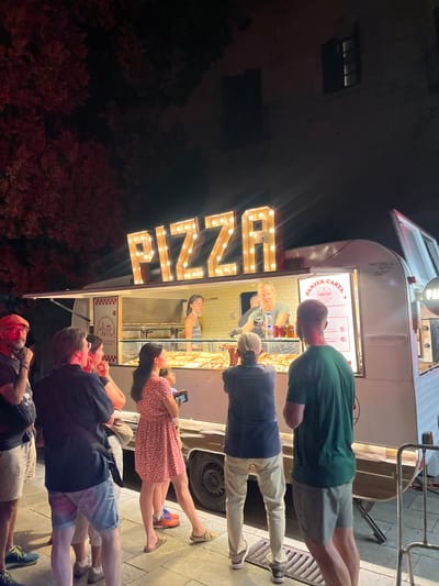 A group of people are gathered outside a food truck named "PIZZA" at night in Sant Cugat del Vallès, Spain. The food truck is illuminated by strings of lights around the "PIZZA" sign. Two workers are visible inside the truck, preparing food. A menu board displays "PANZER CARTA" and other text. Several customers are standing in a queue, looking at the food or waiting to be served. The ground appears to be paved with tiles. Trees and a building are visible in the background.