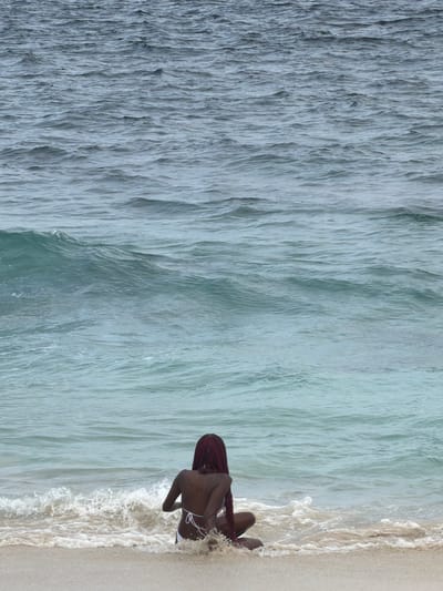 A person with long, dark red hair is sitting on a sandy beach, facing away from the camera and towards the ocean. They are wearing a white bikini top with thin straps. The ocean water is calm with gentle waves lapping at the shore, creating white foam around the person's legs. The sand is light in color. The background consists of the rippling surface of the ocean, with the water changing in color from a lighter turquoise near the shore to a darker blue further out. There are no discernible buildings or landmarks to indicate a specific city location.