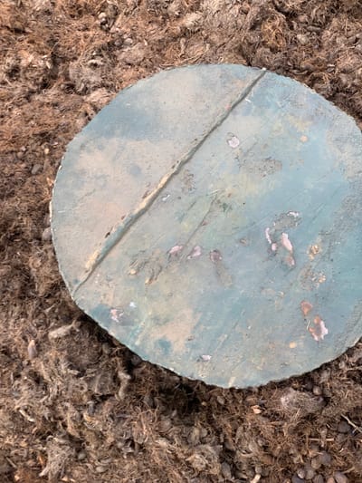 The image shows a circular, weathered, green concrete cover, partially resting on top of a bed of brown, fibrous, mulch. The cover has a large crack running through its center, with a few small, faded orange and pink patches scattered across its surface. There are no visible people, animals, or plants in the image. The location is Egbeda, Nigeria, based on coordinates (latitude: 7.119, longitude: 5.362).