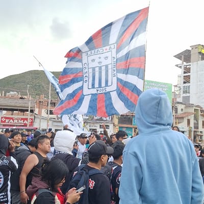 A dense crowd of approximately twenty-five individuals is observed outdoors on a cloudy day in what appears to be a city street or plaza. The scene is located in Wanchaq, Peru.

In the immediate foreground, facing away from the viewer, is an individual wearing a light blue hooded sweatshirt. To their left, a female is seen looking down at a smartphone in her hands. The crowd primarily consists of young adults and men, many wearing casual clothing, including t-shirts, baseball caps, and backpacks. Some individuals are seen holding smartphones, potentially recording or photographing the event.

Dominating the upper center of the image is a large flag, held aloft on a pole. The flag features a red, white, and blue design, with a central emblem consisting of a shield with vertical stripes and three crowns, encircled by text that includes "LA CALLE", "CASCO", and "POOS". The flag's background is a sunburst pattern of alternating red and blue stripes on a white base.

The background reveals an urban environment. On the left, a "Botica FARMA" sign is visible on a building. Further back on the left, a hillside or mountain range is present. On the right, a multi-story building under construction with exposed rebar is visible. Another building to its left displays a large green billboard with Spanish text, partially visible as "CON EL PUEBLO, UN FUTURO MEJOR." A "Corona" sign is also visible in the mid-ground. The overall impression is a gathering, possibly related to a sporting event or public demonstration, given the prominent flag and crowd density.