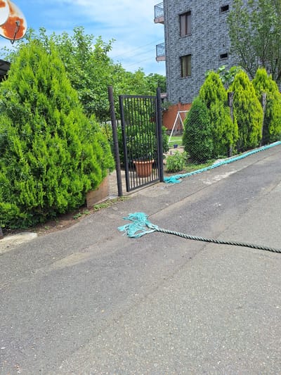 In Kobuleti, Georgia, a road with a slight incline runs past a property with a grey stone building and lush green trees. A black metal gate stands open, revealing a pot and some foliage behind it. A thick, turquoise rope snakes across the asphalt, its frayed ends creating a messy tangle near the gate and stretching further down the road, suggesting it's been laid out or is in use. The scene is under a bright, partly cloudy sky.